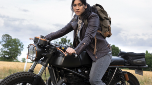 Alaqua Cox is seated on a black Royal Enfield Continental GT 650 motorcycle, parked outdoors with fields in the background and is looking straight into the camera. She is wearing a brown leather jacket, a plaid shirt, dark pants, and has a backpack on.